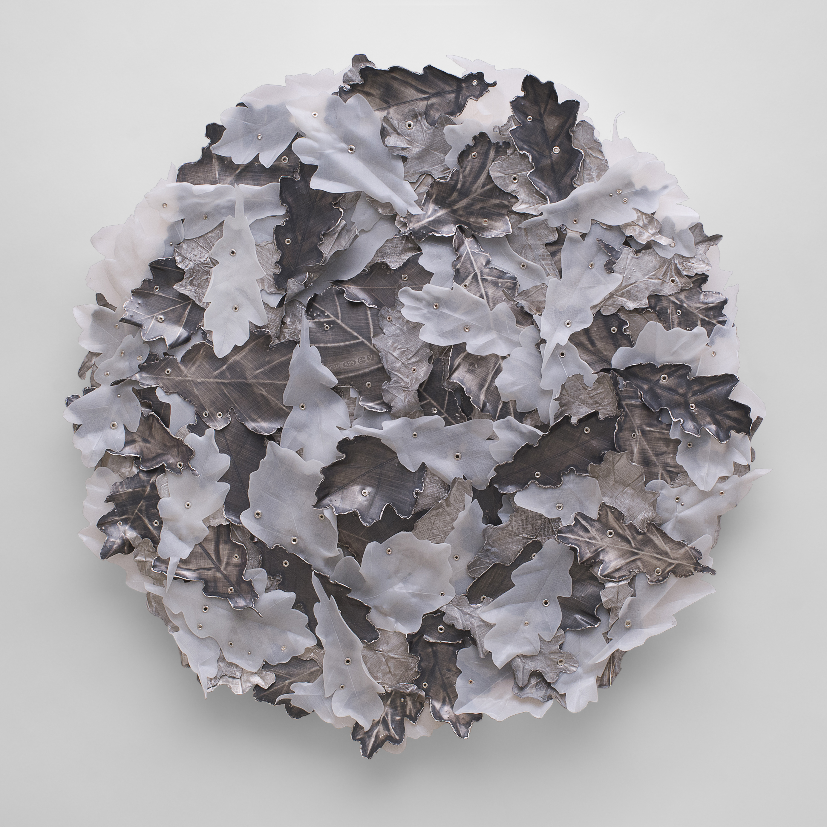 round wall hung artwork with the front randomly covered in layered oak leaves riveted in place made from milky white coloured plastic aged sheet silver and roughed silver aluminium